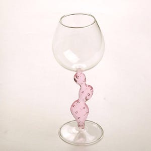May include: A clear glass wine glass with a pink, abstract stem. The stem has a curvy, organic shape with small, round bubbles. The glass has a rounded bowl and a circular base. The overall design is modern and artistic.