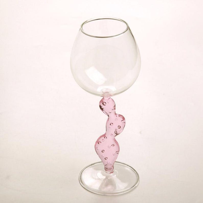 May include: A clear glass wine glass with a pink, abstract stem. The stem has a curvy, organic shape with small, round bubbles. The glass has a rounded bowl and a circular base. The overall design is modern and artistic.