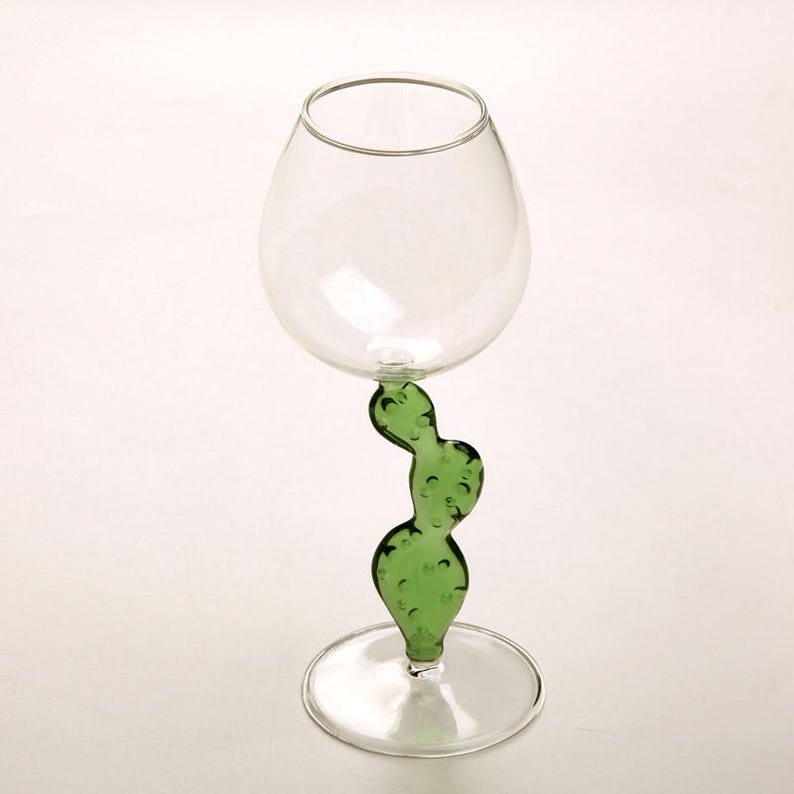 May include: A clear glass wine goblet with a green cactus-shaped stem. The goblet has a rounded bowl and a circular base. The cactus stem is a vibrant green color and adds a unique design element to the glass.