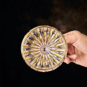 May include: A clear glass coaster with a gold rim and a detailed, sunburst-like pattern. The coaster is held by a hand against a dark background, highlighting the intricate design and the play of light on the glass.