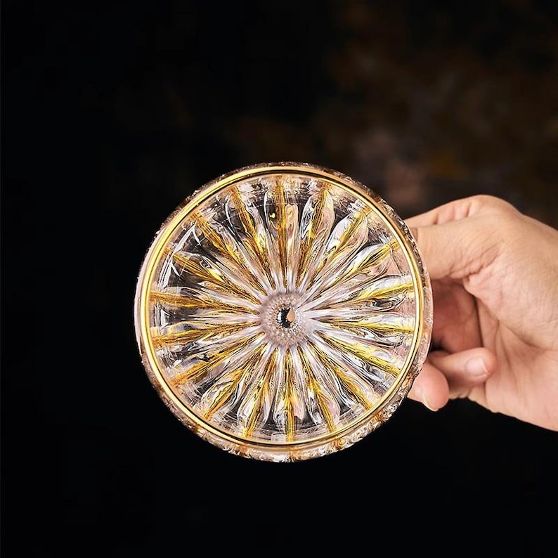 May include: A clear glass coaster with a gold rim and a detailed, sunburst-like pattern. The coaster is held by a hand against a dark background, highlighting the intricate design and the play of light on the glass.