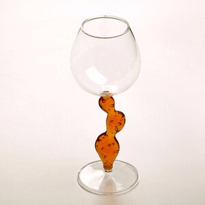 May include: A clear glass wine goblet with a unique stem design. The stem is a curvy, amber-colored shape with small, bubble-like details. The goblet sits on a clear, circular base, and the bowl is round. The background is white.
