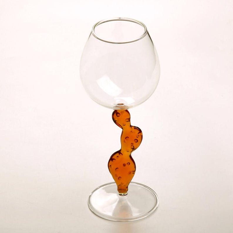 May include: A clear glass wine goblet with a unique stem design. The stem is a curvy, amber-colored shape with small, bubble-like details. The goblet sits on a clear, circular base, and the bowl is round. The background is white.