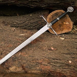 May include: A long, silver sword with a black handle and a decorative pommel rests on a pile of weathered logs. The blade has a series of parallel grooves. The sword's crossguard is silver and has a cross shape.