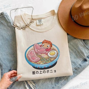 Puede incluir: Camiseta color crema con una ilustración de dibujos animados de un personaje en un cuenco de ramen con carne y un huevo. La camiseta está sobre unos vaqueros azules, con un sombrero marrón y un collar plateado.