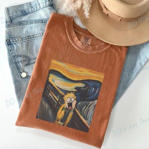 Pode incluir: Camiseta cor de ferrugem com uma paródia da pintura "O Grito" de Edvard Munch. O gráfico retrata uma personagem de desenho animado no estilo da famosa obra de arte. A camiseta está sobre jeans azul claro e um chapéu bege.
