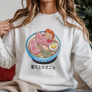May include: White long-sleeve sweatshirt featuring a colorful illustration of a ramen bowl with a cartoon character. The bowl contains noodles, meat, an egg, and green onions. Japanese text is below the bowl.