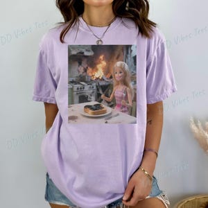 May include: A light purple t-shirt featuring a graphic of a Barbie doll in a pink dress, holding a knife and fork, with a burnt piece of toast on a plate. The background shows a kitchen on fire. Text on the shirt reads "DD Vibes Tees".