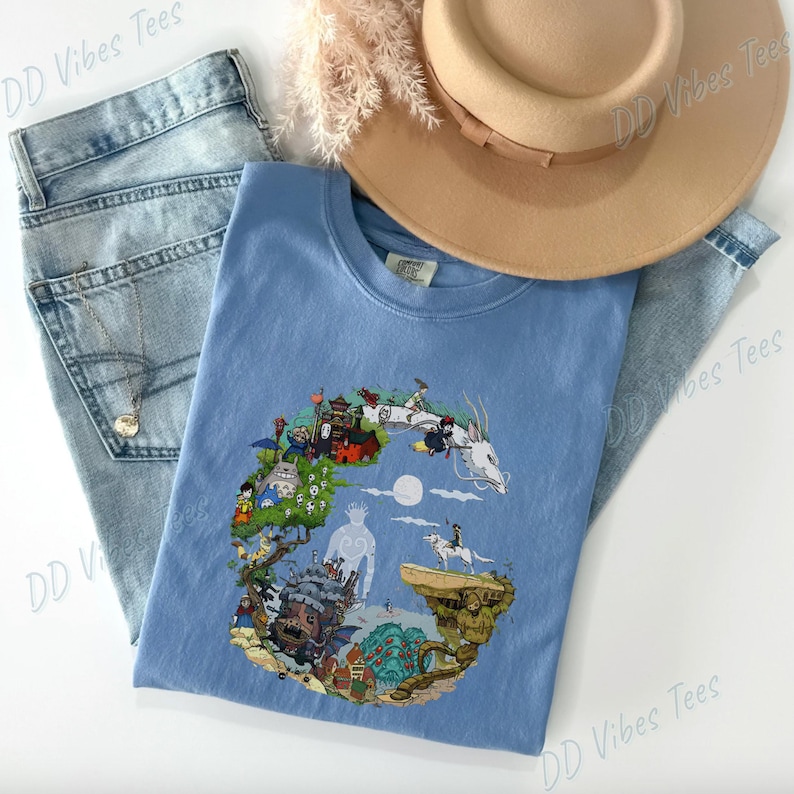 Puede incluir: Una camiseta azul claro con un dise&ntilde;o gr&aacute;fico colorido de un paisaje de fantas&iacute;a con personajes y edificios. La camiseta se combina con vaqueros de mezclilla de lavado claro y un sombrero color canela. El texto "DD Vibes Tees" es visible en el fondo.