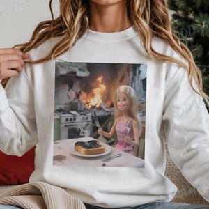 May include: White long-sleeve sweatshirt with a printed image. The image shows a Barbie doll holding a knife and fork, with a slice of burnt toast on a plate. A kitchen fire is in the background.