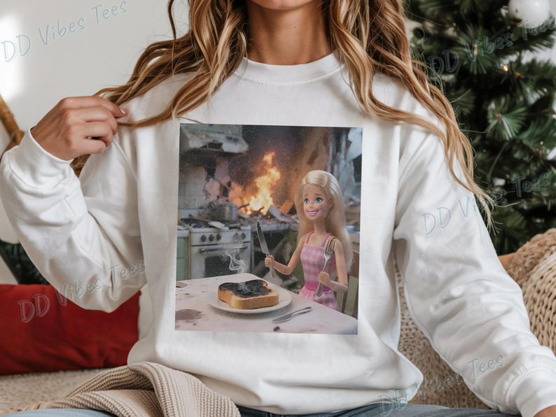 May include: White long-sleeve sweatshirt with a printed image. The image shows a Barbie doll holding a knife and fork, with a slice of burnt toast on a plate. A kitchen fire is in the background.