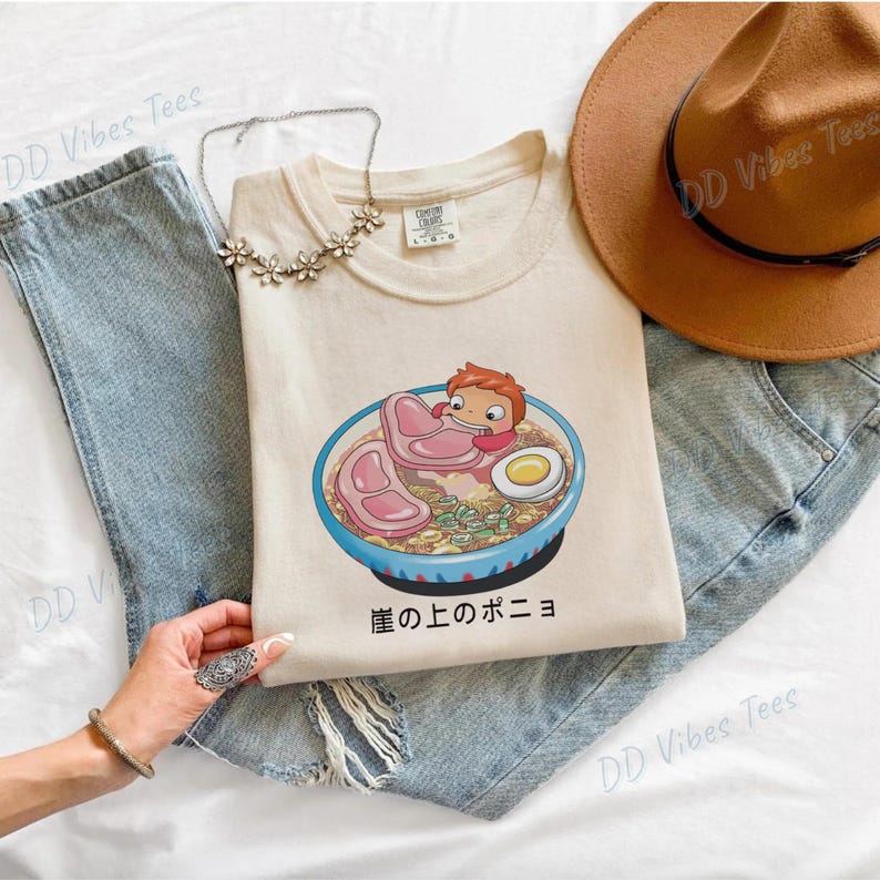 May include: Cream-colored t-shirt featuring a cartoon illustration of a character in a bowl of ramen with meat and an egg. The shirt is laid on top of blue jeans, with a brown hat and a silver necklace.