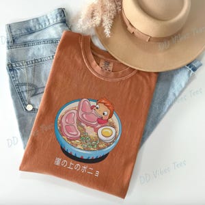 May include: Rust-colored t-shirt featuring a cartoon ramen bowl with a character, meat slices, and an egg. The shirt is paired with light blue jeans and a tan hat. Japanese text is visible on the shirt.