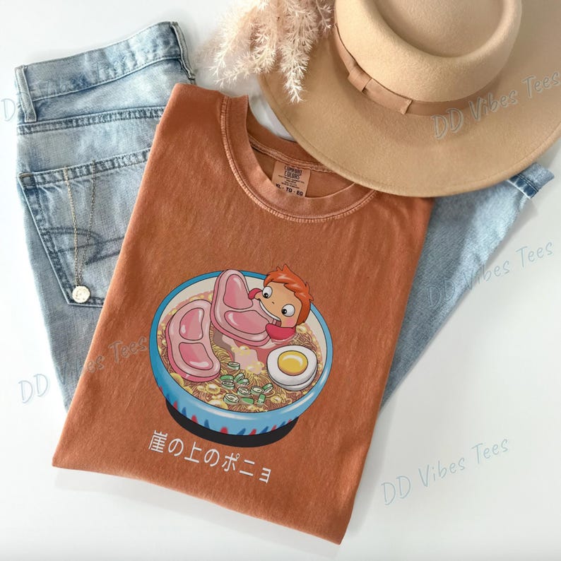May include: Rust-colored t-shirt featuring a cartoon ramen bowl with a character, meat slices, and an egg. The shirt is paired with light blue jeans and a tan hat. Japanese text is visible on the shirt.