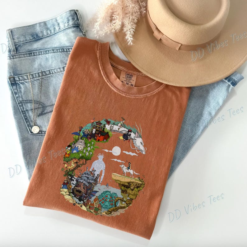 Puede incluir: Camiseta gr&aacute;fica de color &oacute;xido con una colorida ilustraci&oacute;n de personajes y escenas. La camiseta se combina con vaqueros de mezclilla de lavado claro, un sombrero color canela y un collar dorado. El texto "DD Vibes Tees" es visible en el fondo.