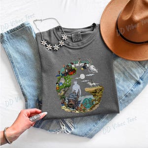Puede incluir: Una camiseta gris con un dise&ntilde;o gr&aacute;fico colorido de un paisaje de fantas&iacute;a con personajes y edificios. La camiseta se combina con unos vaqueros azules, un sombrero marr&oacute;n y un collar floral. El texto "DD Vibes Tees" es visible.