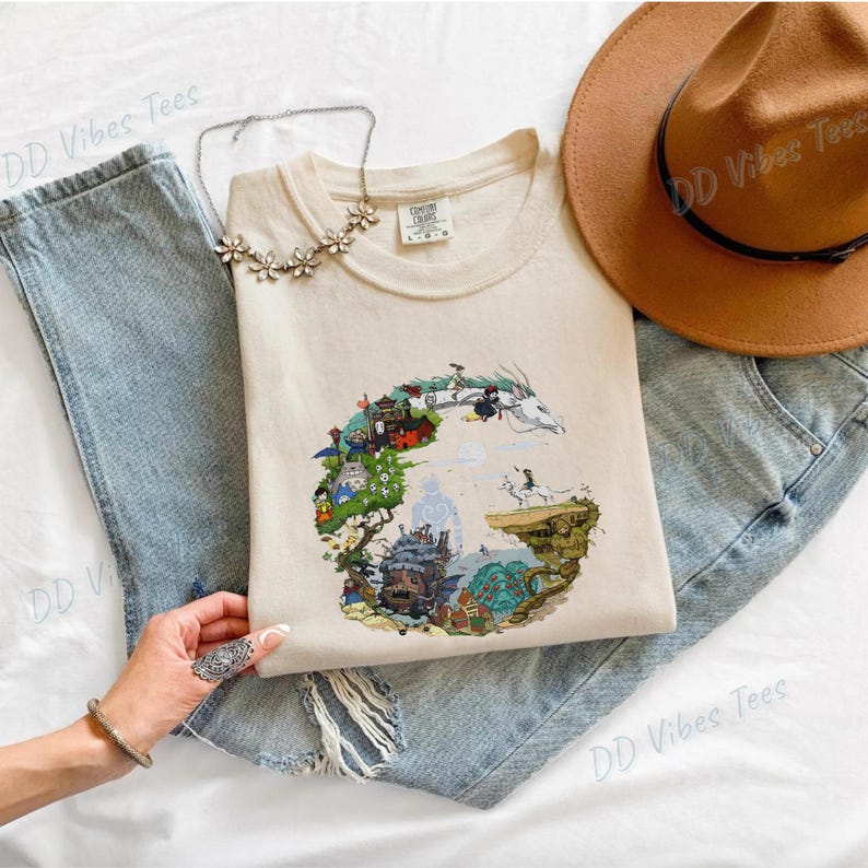 Puede incluir: Camiseta color crema con un dise&ntilde;o gr&aacute;fico colorido de un paisaje de fantas&iacute;a con personajes. La camiseta est&aacute; colocada sobre unos vaqueros azules, con un sombrero marr&oacute;n y un collar plateado. La camiseta tiene el texto "DD Vibes Tees".