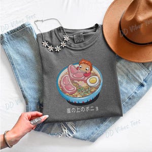 May include: A gray t-shirt featuring a cartoon ramen bowl with a character, ham, and a boiled egg. The Japanese text "崖の上のポニョ" is below the bowl. A silver necklace and a brown hat are also visible.