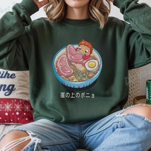 May include: Dark green sweatshirt featuring a colorful illustration of a bowl of ramen with a cartoon character. The ramen includes meat, an egg, and vegetables. Japanese text is below the bowl. The sweatshirt has long sleeves.