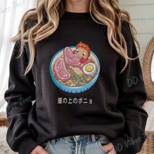 May include: Black sweatshirt featuring a colorful illustration of a bowl of ramen with a cartoon character. The ramen includes pork, an egg, and green onions. Japanese text is below the bowl. The brand "DD Vibes Tees" is printed on the sleeves.