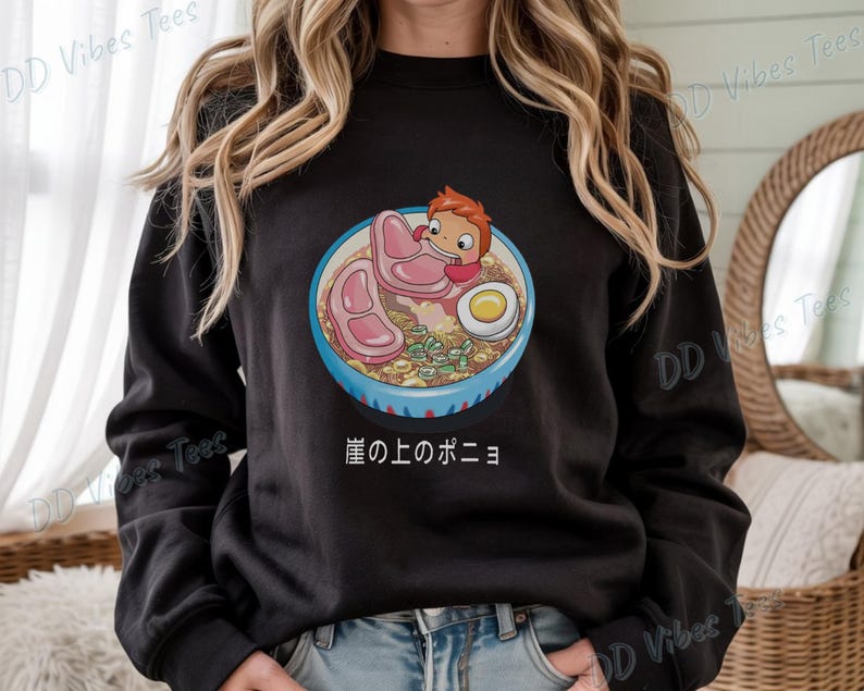 May include: Black sweatshirt featuring a colorful illustration of a bowl of ramen with a cartoon character. The ramen includes pork, an egg, and green onions. Japanese text is below the bowl. The brand "DD Vibes Tees" is printed on the sleeves.