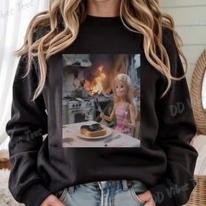 May include: Black long-sleeve sweatshirt with a graphic of a Barbie doll in a pink dress, sitting at a table with burnt toast and a kitchen on fire in the background. The text "DD Vibes Tees" is printed on the sweatshirt.