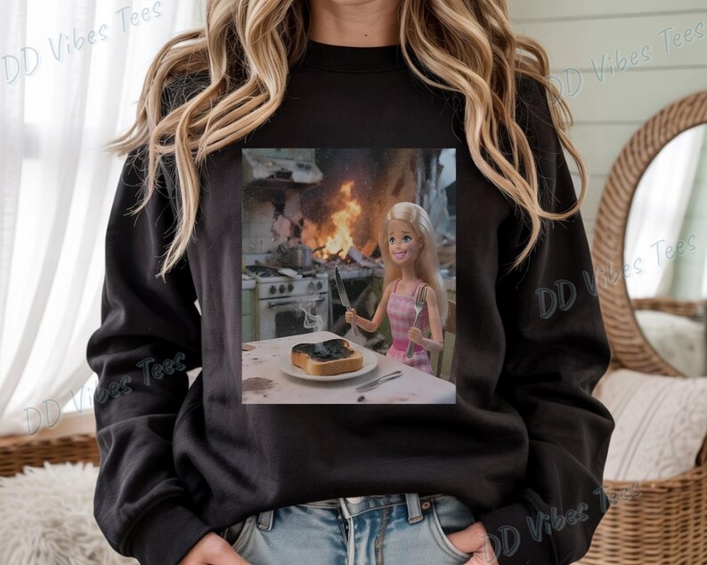 May include: Black long-sleeve sweatshirt with a graphic of a Barbie doll in a pink dress, sitting at a table with burnt toast and a kitchen on fire in the background. The text "DD Vibes Tees" is printed on the sweatshirt.