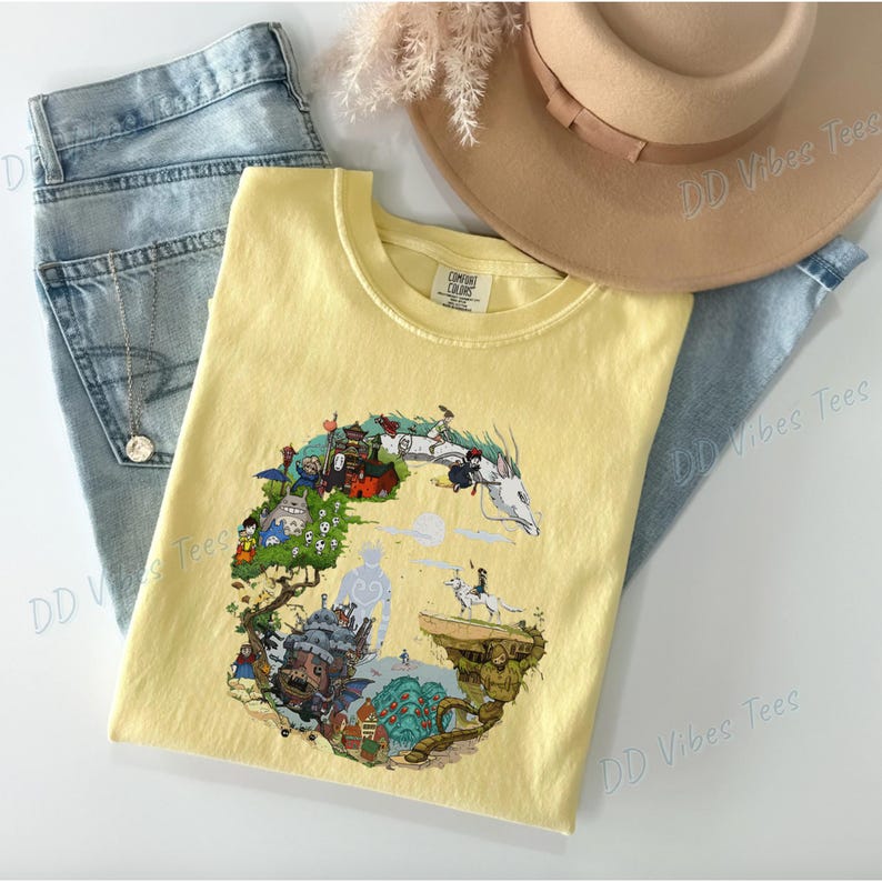 Puede incluir: Una camiseta Comfort Colors de color amarillo claro con un dise&ntilde;o gr&aacute;fico colorido. El gr&aacute;fico representa una escena de fantas&iacute;a con edificios, personajes y un drag&oacute;n blanco. La camiseta se combina con vaqueros azul claro y un sombrero beige.