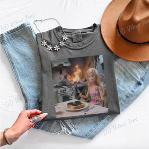 May include: A gray t-shirt featuring a humorous image of a Barbie doll in a pink dress, holding a knife and fork in front of a burning kitchen. The shirt is laid on top of blue jeans, with a necklace and a brown hat.