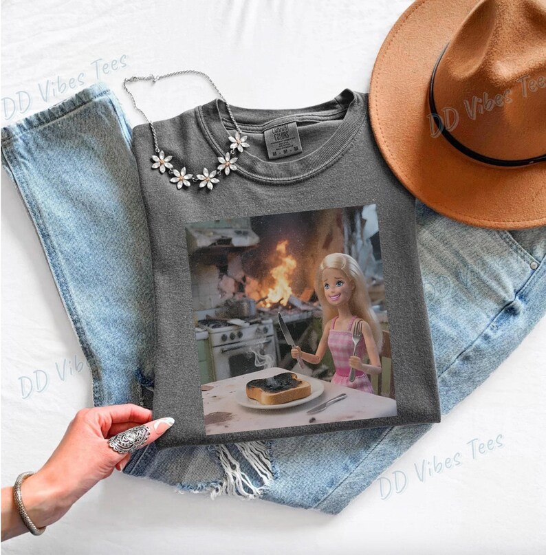 May include: A gray t-shirt featuring a humorous image of a Barbie doll in a pink dress, holding a knife and fork in front of a burning kitchen. The shirt is laid on top of blue jeans, with a necklace and a brown hat.