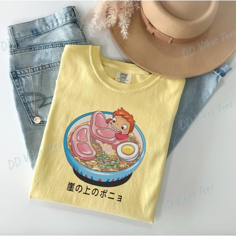 May include: Yellow Comfort Colors t-shirt featuring a cartoon ramen bowl with a character and the Japanese text "崖の上のポニョ". The shirt is laid on top of light blue jeans and next to a tan hat.