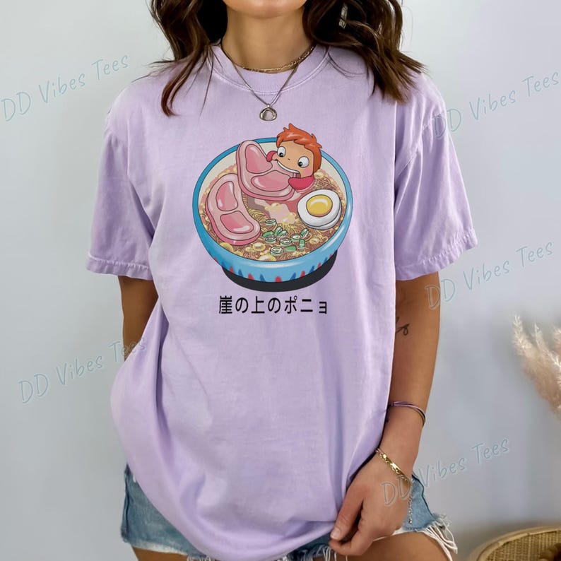 May include: A light purple t-shirt featuring a cartoon illustration of a bowl of ramen with a character from the animated film "Ponyo". The bowl is blue with a white rim and contains noodles, meat, and an egg. Japanese text is below the bowl.
