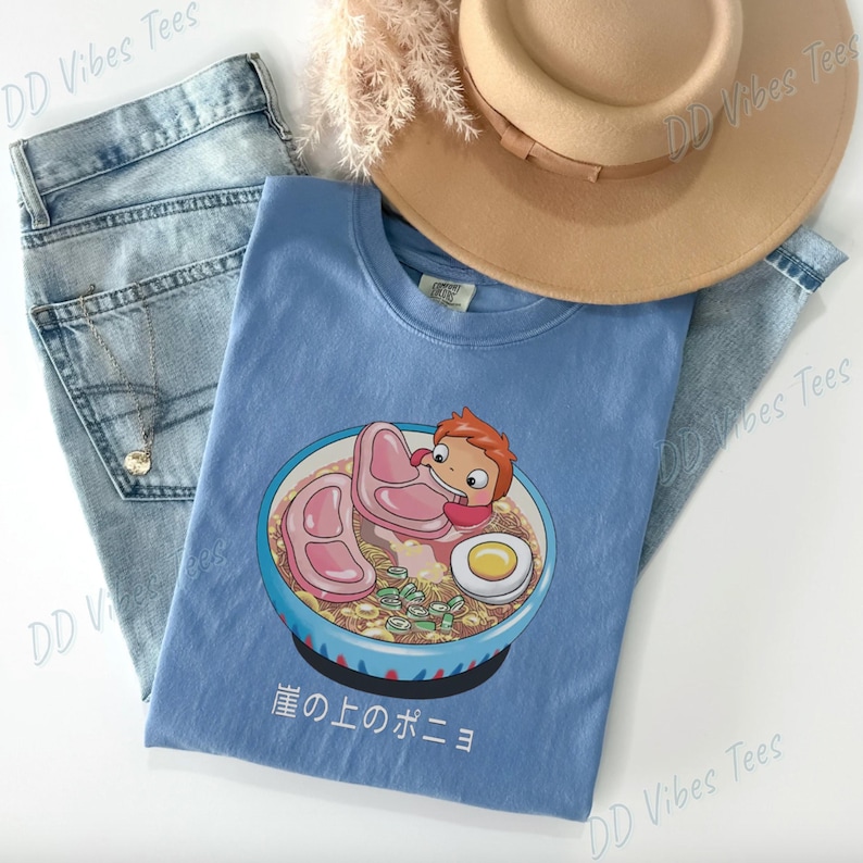 May include: A light blue t-shirt featuring a cartoon illustration of a character in a bowl of ramen with ham and an egg. The Japanese text "崖の上のポニョ" is printed below the bowl. The shirt is laid on top of a pair of blue jeans and a tan hat.
