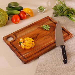 May include: A rectangular wooden chopping board with a groove and a round hole. A stainless steel chef's knife with a black handle rests on the board. Yellow bell pepper slices and fresh vegetables surround the board.
