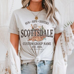 May include: A light gray t-shirt featuring the text SUN SIPS SISTERS SCOTTSDALE CUSTOM GROUP NAME EST. 2026 Desert Weekend Memories. The graphic includes illustrations of a cowboy boot, a disco ball, a cocktail glass, and a cactus.