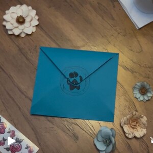 May include: A teal envelope featuring a paw print design. The image also shows paper flowers in shades of white, beige, and blue, arranged on a wooden surface. The envelope is the central focus.