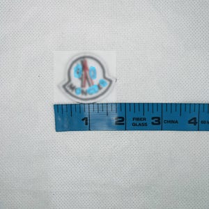 May include: A circular white patch featuring a black and red graphic logo with the text MONCLER, positioned above a blue measuring tape that shows the patch has a diameter of approximately 1.5 inches on the imperial scale.