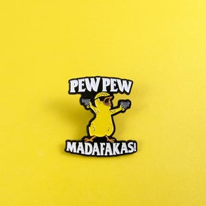Puede incluir: Un pin con forma de pollito amarillo con las palabras "PEW PEW MADAF*KAS!" en letras blancas y negras. El pollito sostiene dos pistolas negras. El fondo es de color amarillo liso.