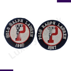 Pode incluir: Dois patches circulares com borda azul marinho e texto branco "POLO RALPH LAUREN". O centro apresenta um "P" vermelho e um fundo branco com estrelas. Um patch tem "1967" e o outro "1961".