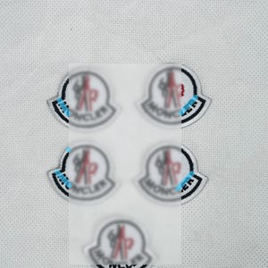 Può includere: Una collezione di cinque toppe ricamate con un emblema circolare, una figura stilizzata, la parola "MONCLER" e un design a scudo. Le toppe sono bianche, nere, rosse e blu. Sono su uno sfondo di tessuto bianco.