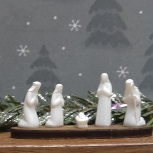 May include: A miniature nativity scene with five white figures and a manger on a wooden base. The background features a gray pattern with snowflakes and Christmas trees, with a garland of green and silver tinsel.