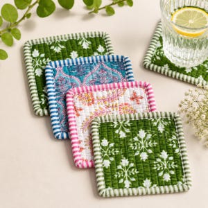 May include: A set of four square quilted fabric coasters featuring diverse patterns. Two coasters have a green and white floral print with green striped edges, one has a blue and white intricate pattern with blue striped edges, and one has a pink and white floral design with pink striped edges.