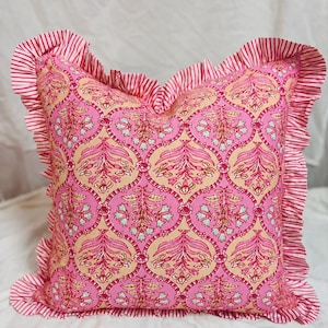 May include: A square throw cushion featuring a repeating pink and cream floral pattern with a central botanical motif. The cushion is bordered by a ruffled edge made of fabric with thin, vertical pink and white stripes that frame the entire item.
