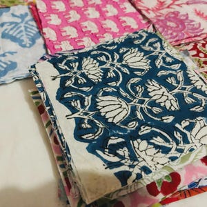 Puede incluir: Una pila de servilletas de tela cuadradas con varios patrones estampados en bloque. La servilleta superior muestra un diseño floral azul oscuro sobre un fondo blanco. Otros patrones visibles incluyen elefantes rosas, hojas azul claro y motivos florales rojos.
