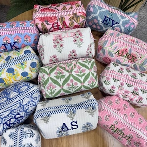 May include: A collection of quilted fabric pouches in various floral patterns and colors, including blue, pink, white, and green. Each rectangular pouch features a zippered closure and is personalized with embroidered initials or names in contrasting thread.