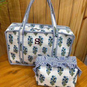 Könnte beinhalten: Eine gesteppte rechteckige Tragetasche und eine passende kleinere Tasche, beide mit weißem Grund, blauem Blumenmuster und blau-weiß gestreiftem Besatz. Die Tragetasche zeigt einen auffälligen, rot gestickten Buchstaben S auf der Vorderseite.