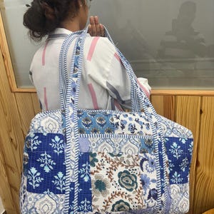 May include: A large rectangular quilted duffel bag featuring a patchwork design of various blue and white floral and paisley patterns. The bag has two long shoulder straps made of a blue and white striped fabric that matches the trim of the bag.