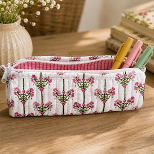 May include: A rectangular quilted pencil case featuring a white fabric base with a repeating pattern of pink flowers and green stems. The interior is lined with pink and white striped fabric, and the case contains three pens in yellow, pink, and green.
