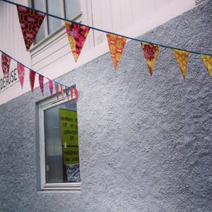 May include: A line of colourful triangular fabric bunting hangs against a textured grey wall. The bunting features various patterns and colours, including floral and geometric designs. A window is visible below the bunting.