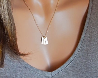 Puede incluir: Un collar de cadena dorada con tres pequeños colgantes rectangulares que cuelgan de él. Los colgantes están hechos de un metal plateado y tienen una textura martillada.